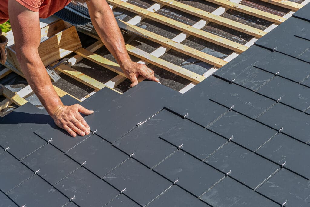 A person installing dark gray slate tiles on a sloped roof, using wooden battens as support. The person is positioned on the left, actively laying the tiles. The roof is partially completed in the foreground.