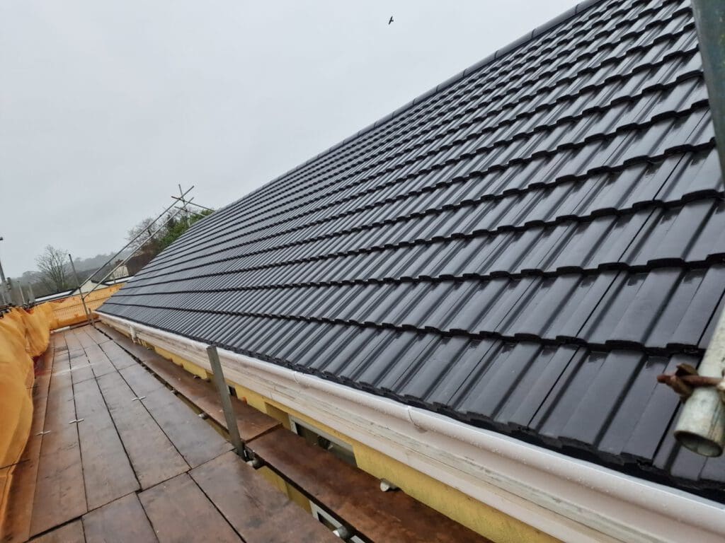 View of a newly installed black tiled roof on a building under overcast skies. The roof is surrounded by a scaffolding platform with wooden planks and protective barriers. A lone bird flies in the distance.