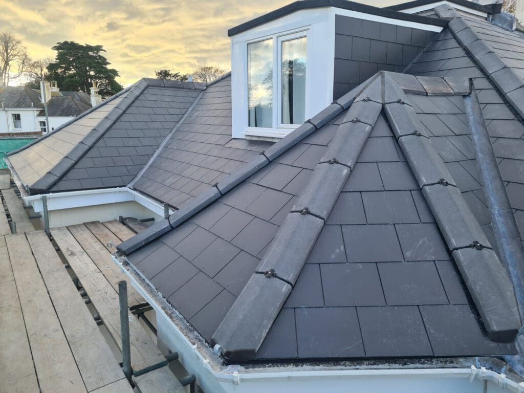 A newly constructed dark slate roof with multiple peaks and a dormer window. The roof is surrounded by scaffolding, and trees are visible in the background under a cloudy sky.