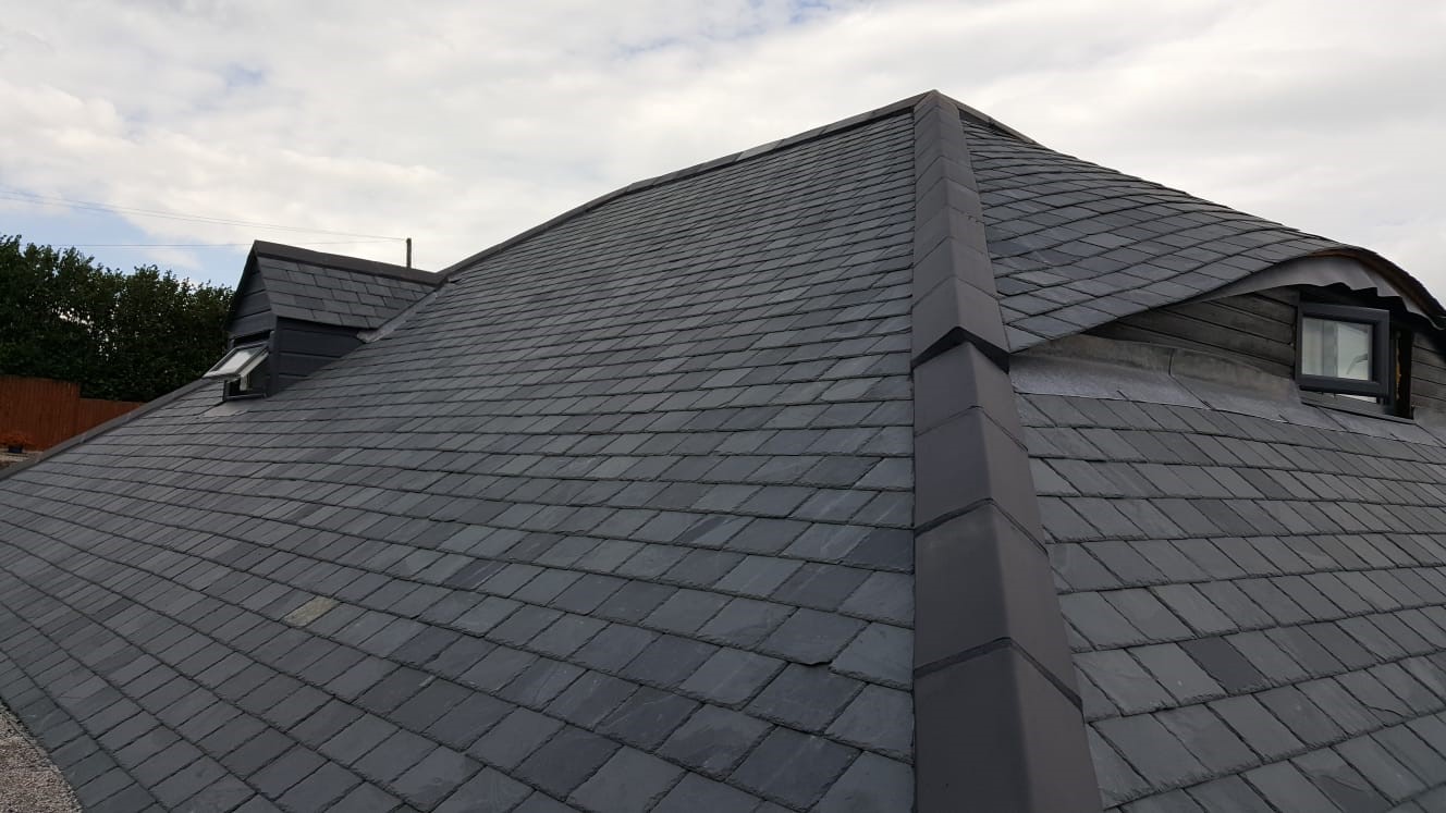 A large slate-tiled roof with two dormer windows under a cloudy sky. The slates are dark gray and arranged in neat rows, creating a modern yet traditional appearance.