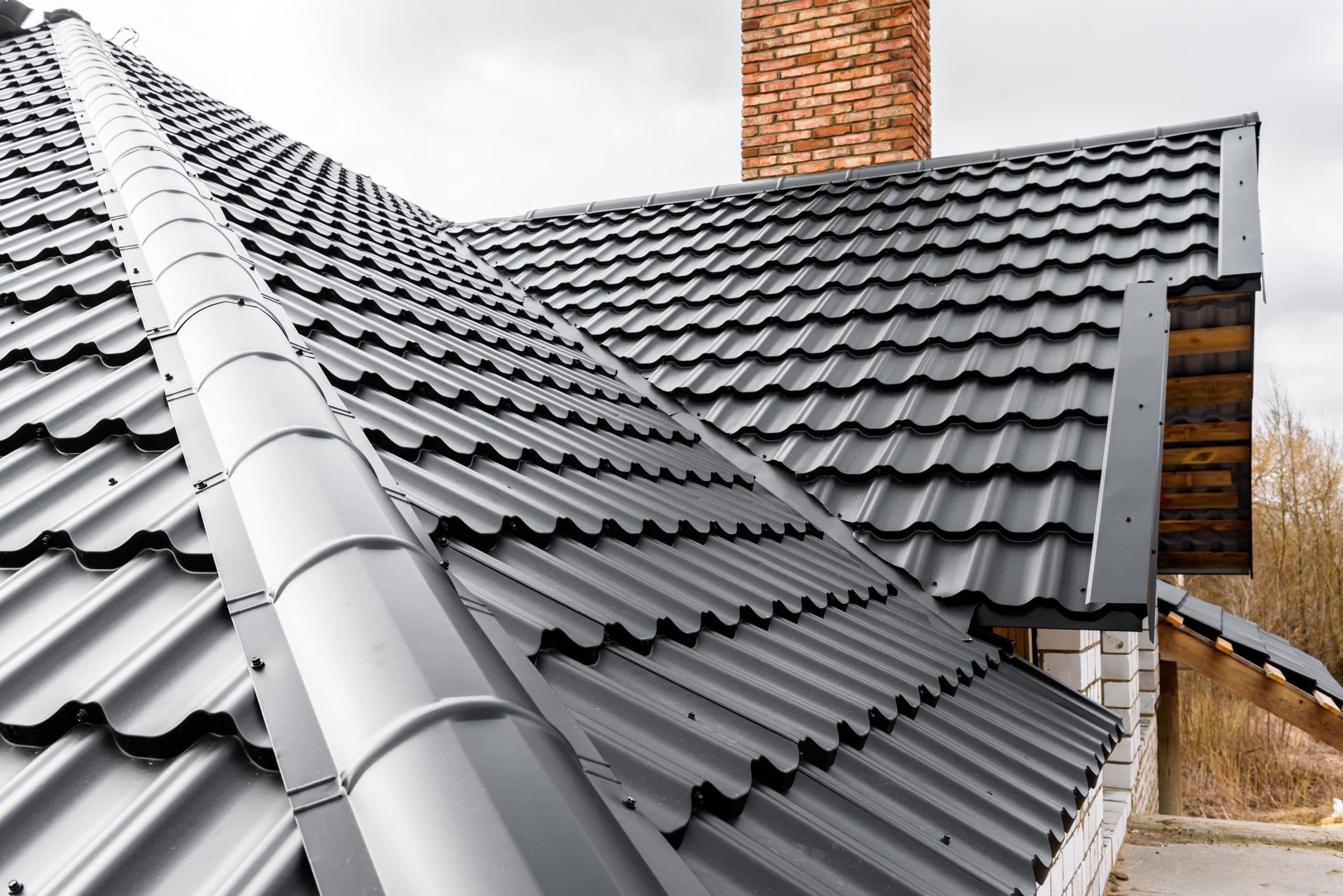 A close-up view of a house roof with dark gray metal tiles, featuring a brick chimney. The roof has a modern, shiny finish and sits against a cloudy sky, with a glimpse of trees in the background.