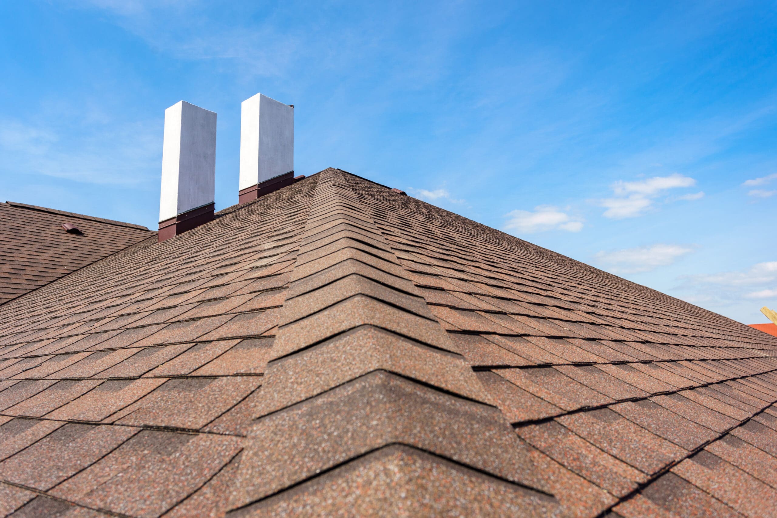 The image shows a rooftop with brown asphalt shingles under a blue sky. Two white chimneys extend vertically from the roof, and a few scattered clouds are visible in the sky.