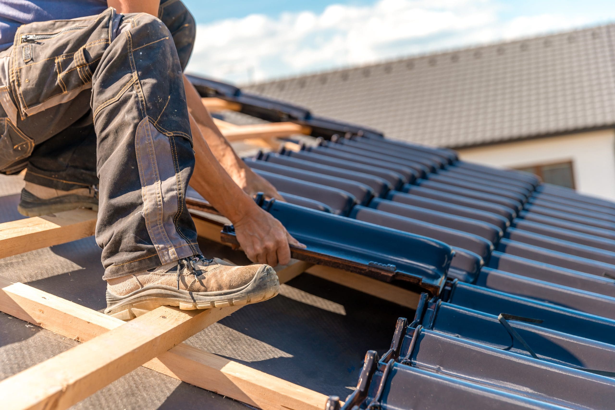 A person in work attire installs dark roof tiles on a sloped wooden frame under a clear sky. The individual is kneeling, with their hands adjusting a tile. A finished section of the roof and part of a building are visible in the background.