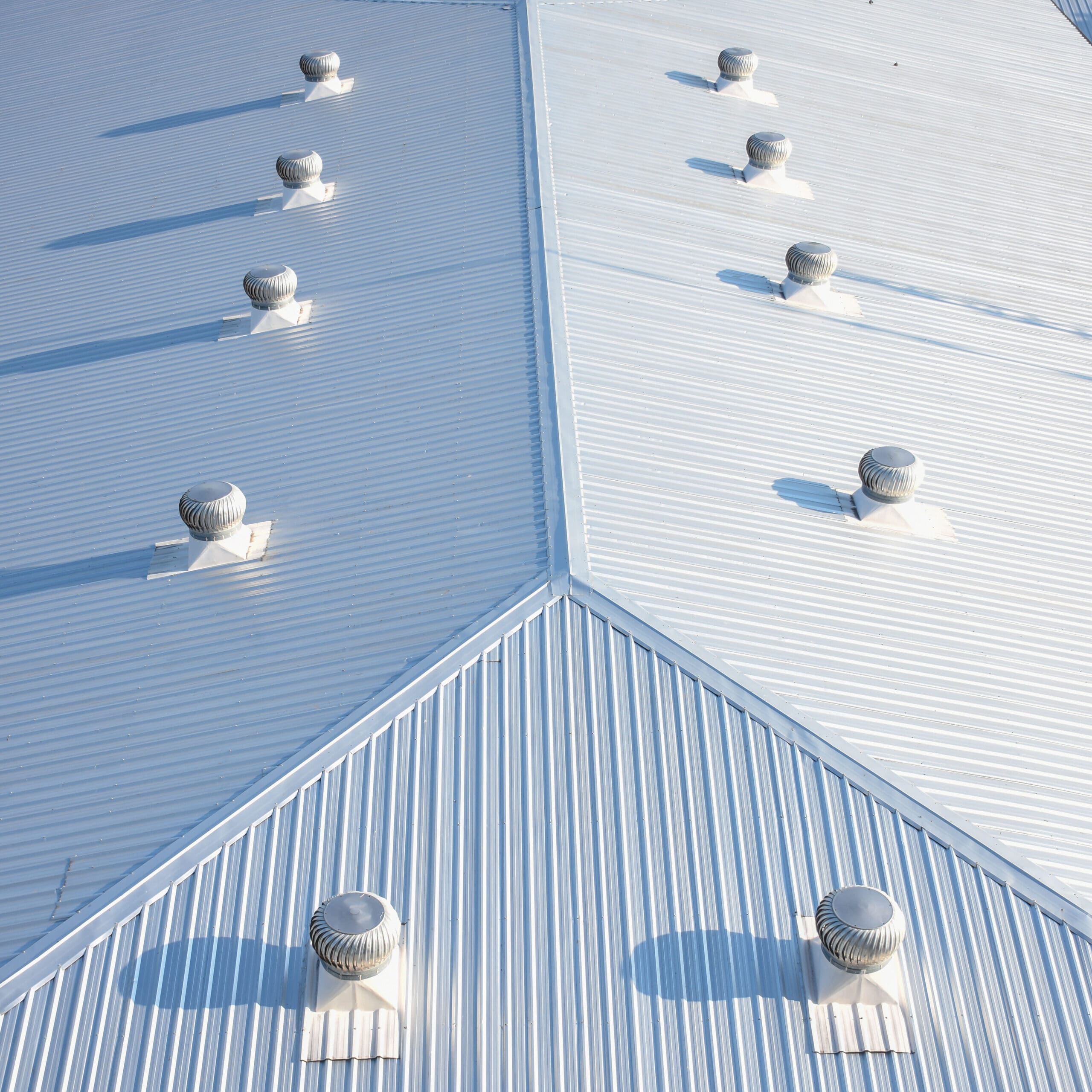 Aerial view of a metallic rooftop with parallel ridges. Eight evenly spaced round ventilation fans are aligned in two diagonal rows, casting long shadows across the surface, creating a pattern of light and shade.