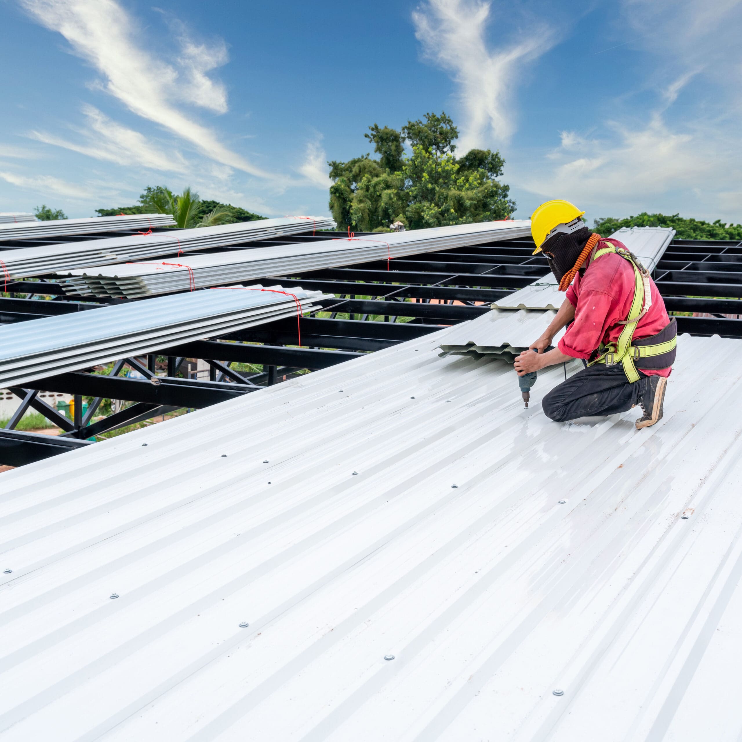 A construction worker wearing a yellow hard hat and safety harness is kneeling on a metal roof, securing panels with a power drill. The sky is clear with wispy clouds, and trees are visible in the background.