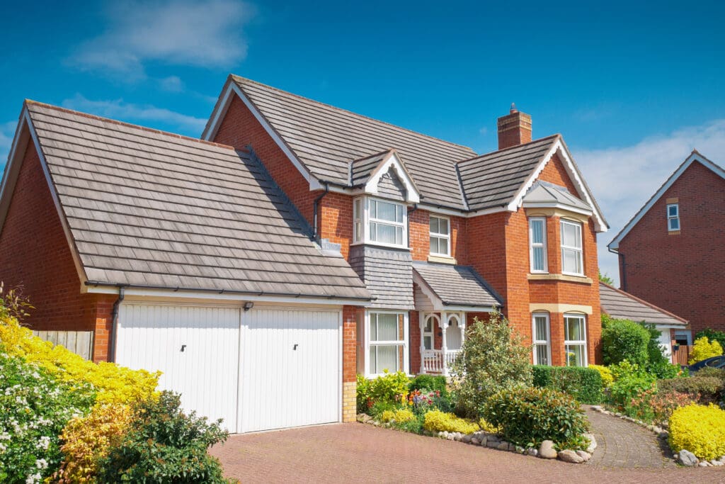 A two-story red brick house with a tiled roof, featuring a double garage and a garden with various shrubs and flowers. The sky is clear and blue, enhancing the vibrant appearance of the home and landscaping.