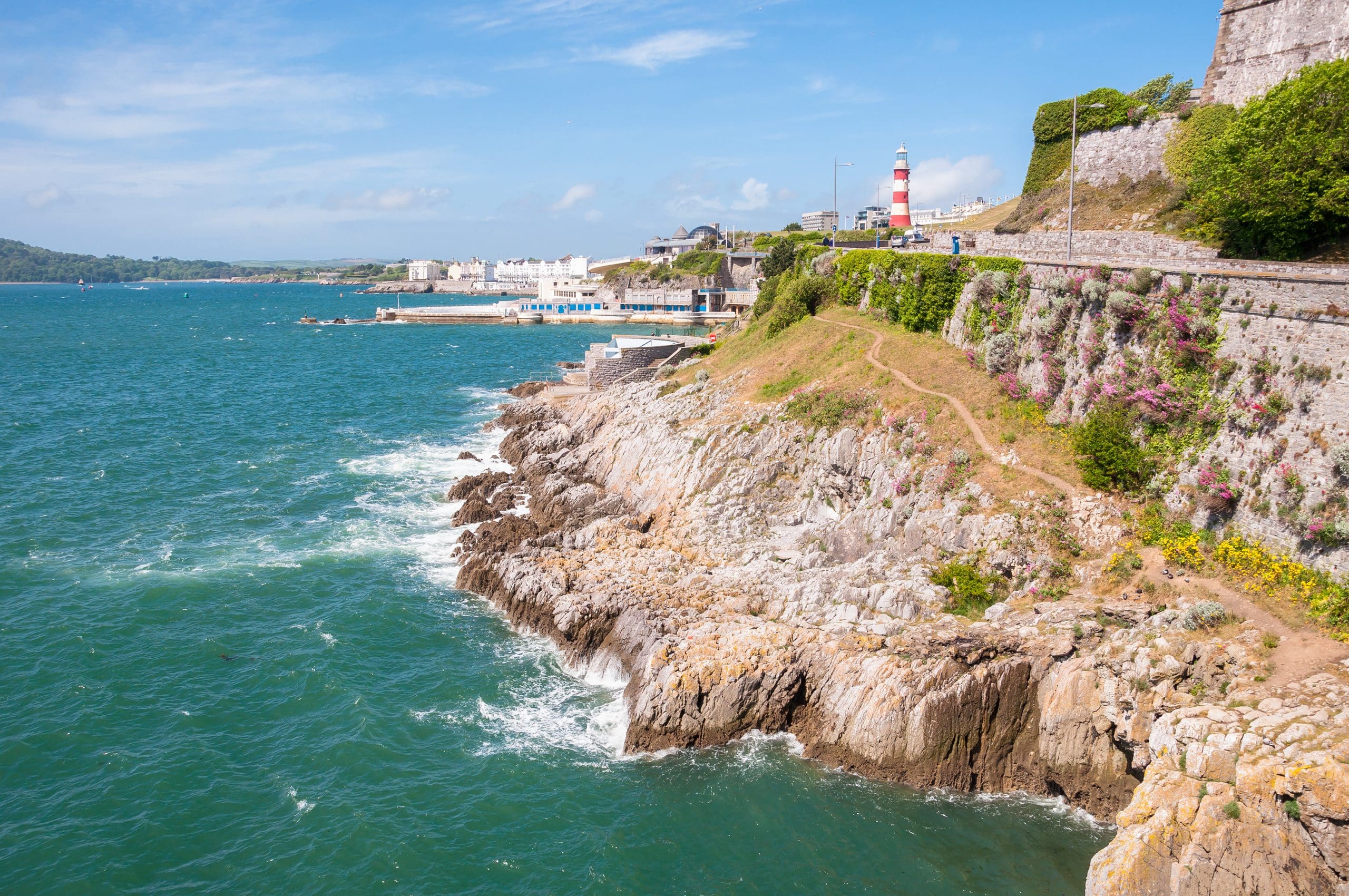 Coastal scene with rocky cliffs and waves, featuring a red and white striped lighthouse. A walking path winds along the cliffs under a clear blue sky, with greenery and flowers adorning the area. Distant buildings are visible along the shoreline.